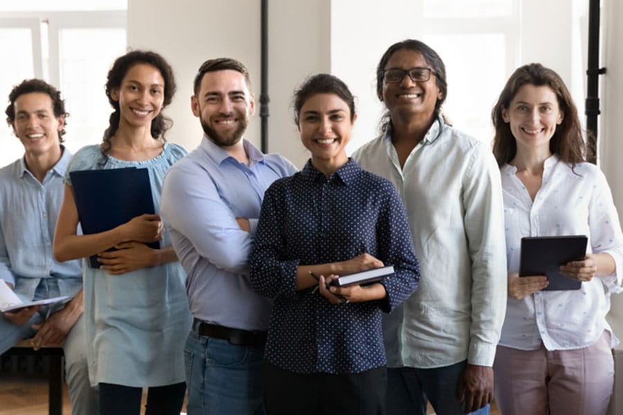 A diverse group of six smiling adults, some holding notebooks or tablets, stand close together indoors, confidently looking at the camera—perfectly capturing the spirit of Metro Detroit ESL’s Fall Registration and Semester Update.