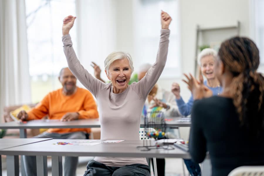 A joyful elderly woman raises her arms in celebration while playing bingo, one of many fun ESL activities enjoyed by all ages in a bright, cheerful room. Others around her are smiling and clapping, sharing in her excitement.