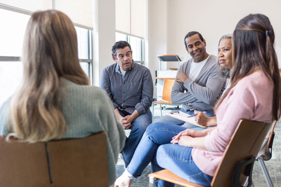 Five adults sit in a circle in a bright room in Metro Detroit, engaged in an ESL group discussion. Three people are facing forward, listening intently, while two others are seen from behind.