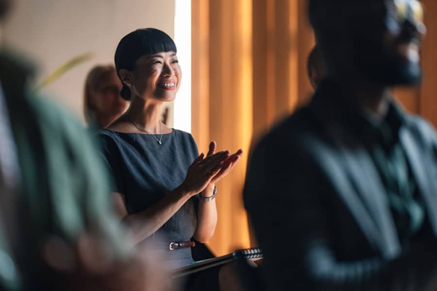 A woman in a dark dress is smiling and clapping while seated among others in a well-lit room, suggesting she is attending an event or presentation featured in the latest ESL News from Metro Detroit.