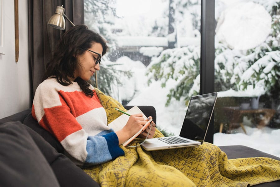A woman sits on a couch with a colorful sweater and a yellow blanket, writing in a notebook while using a laptop. Snow-covered trees are visible outside the window as she explores easy ways to practice English.