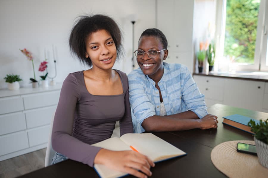 Two women sit at a table indoors, smiling at the camera as they discuss fun ways to practice English. One wears glasses and a blue plaid shirt, while the other sits by an open notebook in a bright room filled with plants and decor.