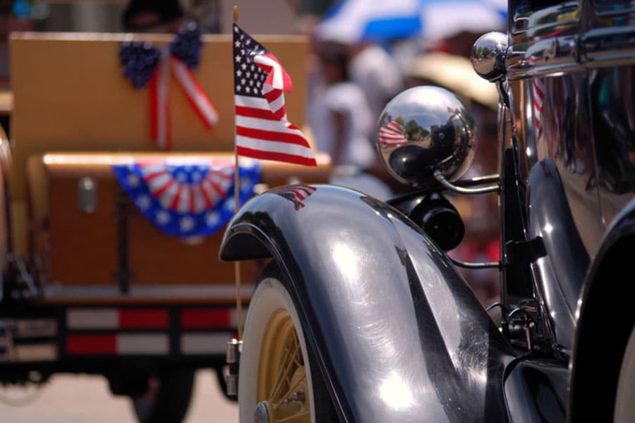 Close-up of a vintage car with an American flag on the fender during an Independence Day parade. In the background, another vehicle is decorated with patriotic red, white, and blue bunting and bows. People are blurred in the distance.