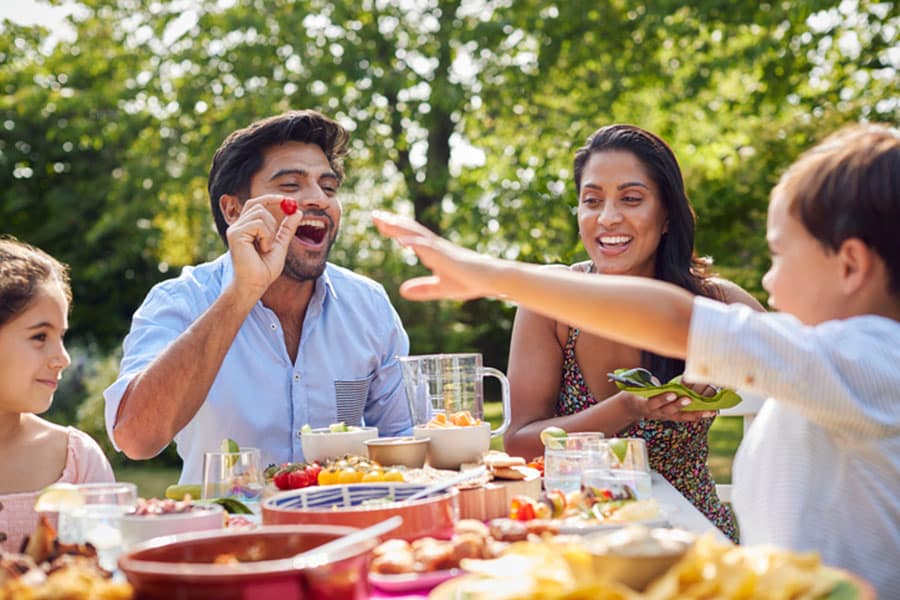 A family sits at an outdoor table enjoying a meal together on Independence Day. A man playfully holds up a cherry tomato while smiling at a child reaching for food, with the table full of festive dishes under bright daylight.