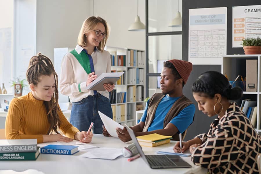 Four people sit at a table in a classroom in Sterling Heights, working with papers, notebooks, and a laptop. A standing woman with a tablet observes them, while English learning materials and charts—typical of ESL classes—are visible in the background.
