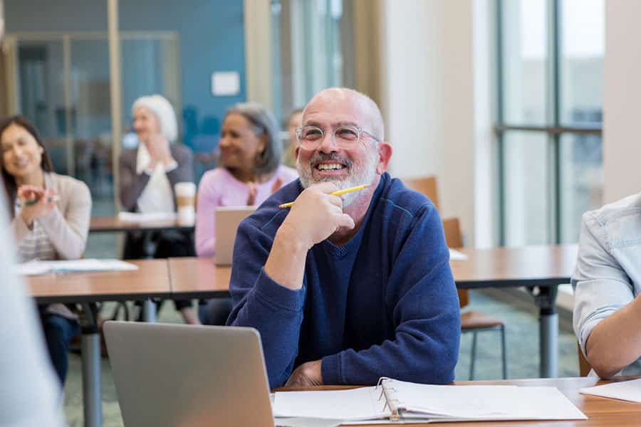 An older man with glasses smiles and holds a pencil during an ESL class session. Other adults sit at desks, listening and engaging in the background as sunlight streams in through large windows.