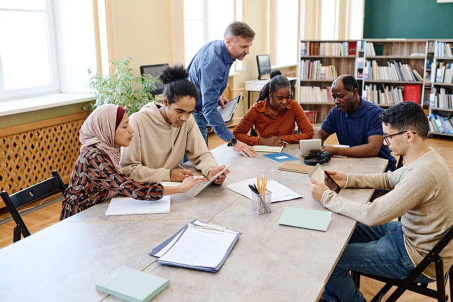 A diverse group of students sits around a table in a library in Clawson, Michigan, working together with notebooks and tablets during their English classes, while a teacher stands and assists them. Bookshelves and windows are visible in the background.