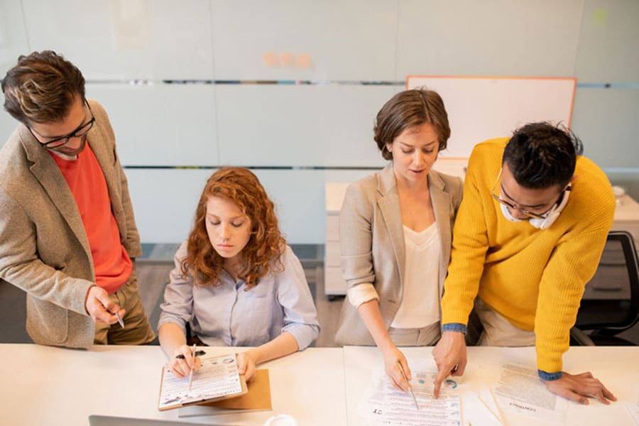 Four people work together at a desk in an office setting, looking at and discussing documents related to English classes. One person writes notes while others point at papers, all appearing focused and collaborative—just the kind of teamwork you’ll find in successful ESL classes in Clawson, Michigan.