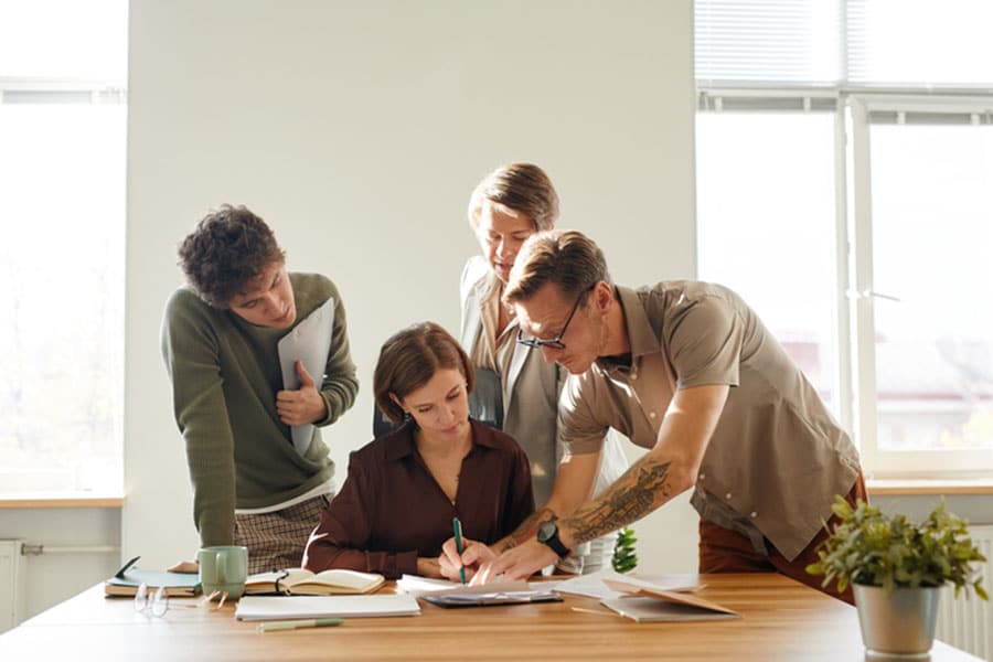 Four people gather around a table with papers and notebooks, collaborating on work. One person writes while others look on and discuss, sunlight coming through the windows behind them. This lively scene could easily take place in one of the ESL English classes held in Sterling Heights, where students come together to learn and grow in a supportive environment.