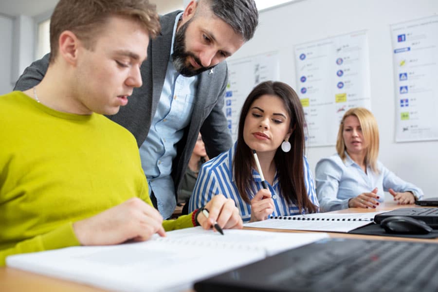 A teacher leans over to help two students with notebooks in an English class in Sterling Heights, while another student sits in the background.