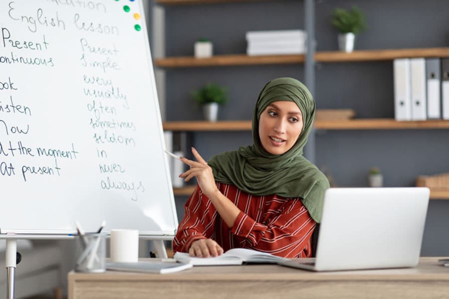 A woman in a green hijab teaches an English lesson online, pointing at a whiteboard with verbs written on it, while sitting at a desk with a laptop and books—an ideal scene for those seeking Free ESL Classes or looking for English Classes Near Troy.